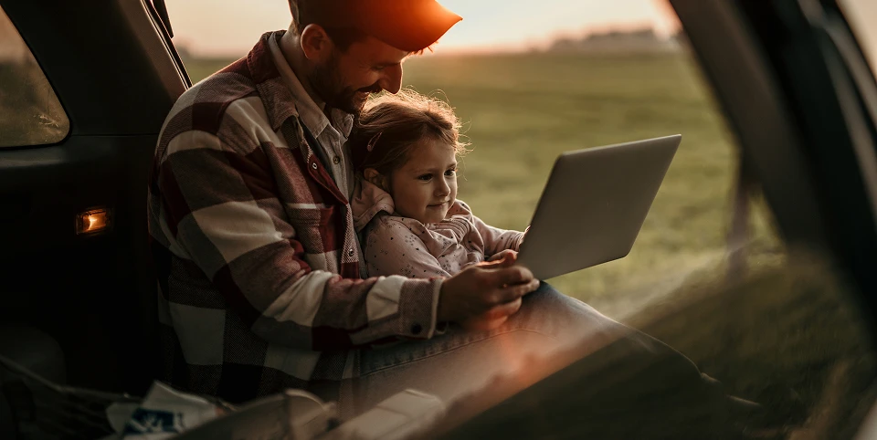 Un tată și copilul lui privesc un laptop într-o mașină parcată